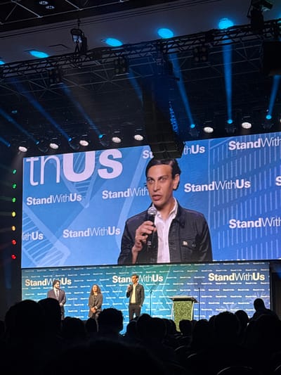 An indoor event is captured within a large conference hall or ballroom in Las Vegas, United States. The scene features a brightly lit stage dominated by a large LED screen displaying a blue background with the prominent white text "thus" and repeated instances of "StandWithUs." Subtle architectural outlines of buildings are visible within the screen's graphic design. Above the screen, a dark truss system supports multiple stage lights emitting visible blue light beams and suspended line array speakers.
On the stage, three individuals are visible. A male speaker, centrally positioned, has dark, slicked-back hair, is wearing a white collared shirt and a dark jacket, and is speaking into a microphone held in his right hand. His enlarged image is simultaneously displayed on the overhead LED screen. To his left, a male in a dark suit with a light shirt and tie stands attentively. To the main speaker's right, a female with dark hair and glasses, wearing a dark blazer, also stands facing the audience. A dark podium with a textured green and gold front is situated to the right of the main speaker.
The foreground is occupied by the dark silhouettes of numerous seated audience members, indicating a large gathering attending the event, with their heads and upper torsos visible against the illuminated stage.