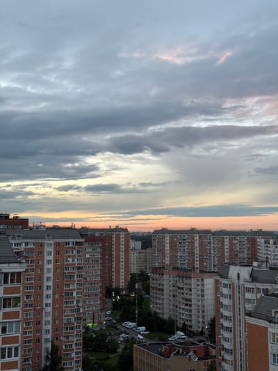 This media file depicts a cityscape at dusk in Otradnoye, Russia. The scene is dominated by a dramatic sky filled with a mix of dark, brooding clouds and streaks of lighter, illuminated clouds tinged with pink and orange, suggesting the last rays of sunlight. Below the sky, a dense collection of apartment buildings stretches across the frame.
The buildings are primarily multi-story structures with many windows, indicative of residential housing. Some buildings feature brick facades in shades of orange and brown, while others are clad in lighter materials. The architecture appears to be of a Soviet-era or post-Soviet style, common in many Russian cities. In the background, glimpses of more buildings and trees can be seen, along with parked cars and streetlights, hinting at an active urban environment.
The time of day is clearly indicated by the sunset colors in the sky and the artificial lights beginning to illuminate the city. The weather appears to be partly cloudy, with the clouds creating a dynamic and somewhat somber atmosphere. There is no visible text in the image. The overall impression is one of urban life transitioning into the evening under a striking sky.