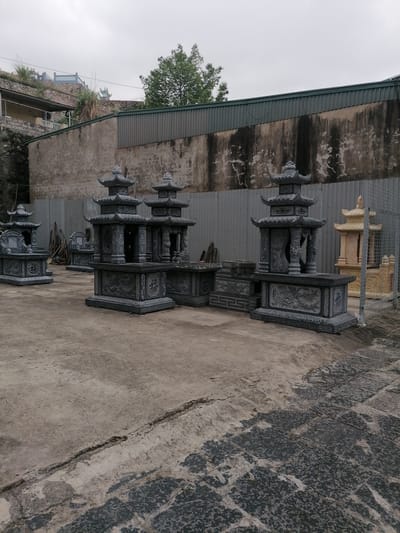 The image depicts a display of several traditional Asian tomb-like structures or memorials in what appears to be a sales or production yard. The structures are made of stone, primarily a dark grey, with intricate carvings. There are multiple pagodas and pedestals arranged on a weathered concrete surface. The setting seems to be outdoors, evidenced by the overcast sky and the presence of a building and wall in the background. The location is identified as Ha Tu Ward, Vietnam. The background consists of a weathered stone wall and corrugated metal fence, with some greenery visible above. There is a scattering of construction materials, pieces of wood and a wire cage.