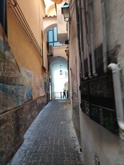 This is a street-level, eye-level, wide-angle photograph of a narrow, paved alleyway in Salerno, Italy. The alley is flanked by buildings with textured walls. The left wall features a mural depicting a coastal town with buildings and water. The right wall has several vertical pipes and is covered in graffiti. In the distance, the alley curves and leads to an archway where a person is visible standing. An air conditioning unit is mounted on the left wall below a window. A street lamp with a blue light is attached to the top of the right wall. The ground is paved with cobblestones.