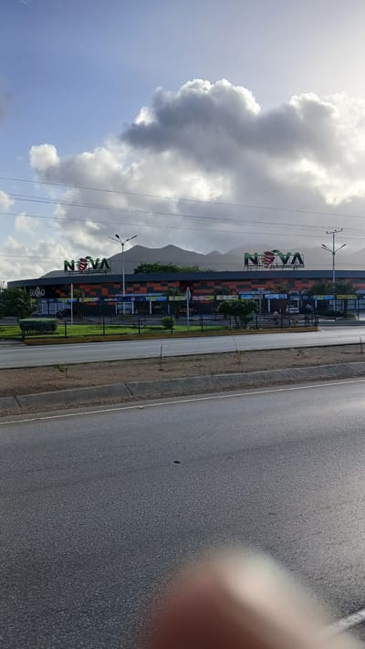The image shows a wide shot of the exterior of the "Nova" supermarket in El Dátil, Venezuela. The building has a modern facade with orange and black checkered patterns and is crowned with large, illuminated "NOVA" signage. Several smaller signs and shop entrances are visible along the front of the building, including "GUJAO". A paved road runs in front of the supermarket, separated by a grassy median with a fence. In the background, mountains are visible under a partly cloudy sky. There are streetlights and power lines in the scene. A few palm trees and other vegetation are planted in front of the supermarket. The image is taken from a slightly elevated position, likely from across the road, with a blurred finger visible in the foreground, suggesting it was taken by hand. No people are clearly discernible in the image.