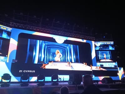 The image depicts a Eurovision performance, possibly during rehearsals or a live show, in San Bartolomé de Tirajana, Spain. A large screen displays the performance with the artist on stage. A smaller screen to the side also shows the performance. The number '21' and the word "CYPRUS" are visible on the screen. Stage lighting is present and there are concert speakers on the stage. The trusses for lighting are visible. Audience members are in the foreground.
