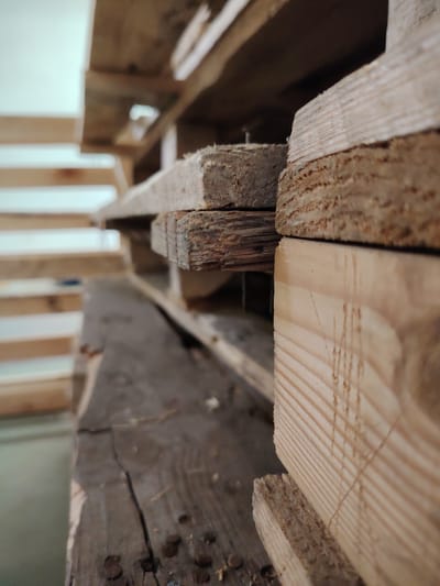 A close-up, low-angle, shallow depth-of-field shot of stacked wooden pallets. The focus is on the textures and grain of the wood, with some pieces appearing weathered and others more freshly cut. Nails are visible on one of the lower, darker wooden beams. The background is blurred, showing what appear to be more wooden structures or shelving, and a light-colored wall. This image was taken at a storage facility or workshop in Domodedovo, Russia. There are no people or discernible activity in the frame.