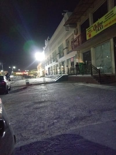The image shows a nighttime view of a building in Termez, Uzbekistan, likely a commercial or office building with a prominent yellow sign advertising "G'n line TAXI". The building features multiple stories with large windows, some of which are illuminated. A walkway or sidewalk runs in front of the building, and a metal railing lines the edge. Several streetlights are visible, casting bright illumination on the scene, and creating lens flare. The ground appears to be paved and wet, suggesting recent rain or a damp environment. In the foreground, a dark-colored car is partially visible on the left side. A few individuals are present, including one person standing near the building entrance and another near the railing. The overall impression is of an urban street scene at night.