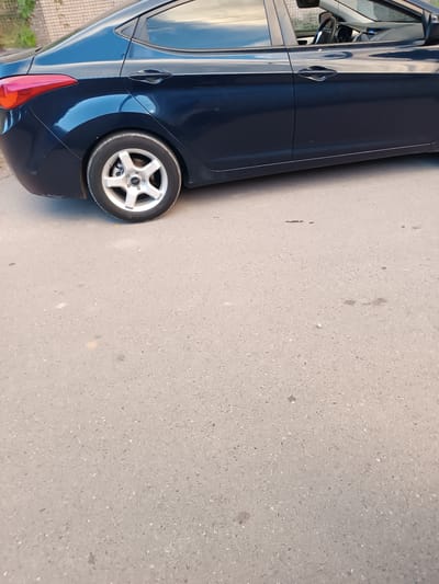 The image shows a close-up side view of a dark blue sedan parked on an asphalt road. The rear wheel and fender of the car are visible on the left side of the frame, along with a portion of the rear bumper and taillight assembly. The car's doors and windows are also visible. The background shows a brick wall and some greenery. The road surface is textured asphalt with some scattered debris. The scene appears to be located in the city of Talin, Armenia. There are no people or any actions depicted in the image.