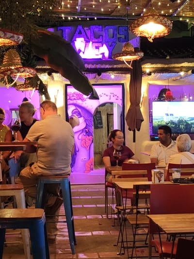 The image depicts a lively outdoor dining scene, possibly a Mexican-themed restaurant or bar. Neon signs illuminate the space, and sombreros are used as decorative lampshades. Several people are seated at tables, some with wine glasses in hand, appearing to be enjoying their meals and conversations. The setting is illuminated with vibrant purple lighting and string lights, creating a festive and energetic atmosphere. Based on the neon sign "TACOS WEY" and the overall decor, the location is likely a popular spot for dining and socializing. There is no evidence that the location is Rotes, Spain.