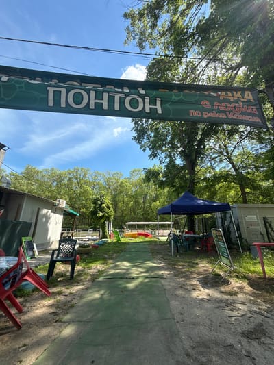 The image shows an outdoor area in Novo Oriahovo, Bulgaria. A green banner with white Cyrillic text is prominent, advertising "Ponton." The banner also contains the words "lodka s lodki po reka Kamzia." Adjacent to the banner is a dirt path leading towards trees. On the right of the path is a green artificial surface. Chairs and tables are scattered around, some under a blue canopy. There are buildings in the background. The sky is blue with light clouds.