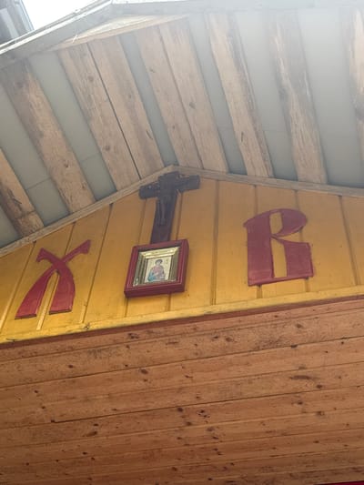 The image shows a close-up view of a yellow wooden wall with religious symbols. On the left side, there is a large red, stylized letter "X". To the right of it, a framed icon of a person is hanging, and above it, a wooden crucifix with a figure of Jesus. To the right of the icon, a large red letter "R" is mounted on the wall. The wooden wall is part of a building's exterior, with visible roof beams and wooden planks overhead. The lower portion of the image displays more wooden planks, likely part of the building's facade. The lighting suggests it is daytime, with sunlight filtering from the top. The overall scene suggests a religious or cultural structure in a rural or historical setting. This specific detail is located in Nenimyaki, Russia.