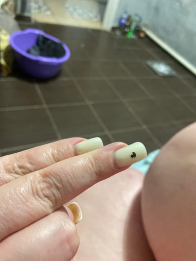 The image shows a close-up of a person's hand, with artificial fingernails. One fingernail has a small black heart design on it. In the background, there is a purple basin and tiled flooring. The environment appears to be indoors, possibly a bathroom or laundry area, with some blurry objects and bottles visible in the distance. There is no indication that this scene is located in the city of Rozkvit, Ukraine.
