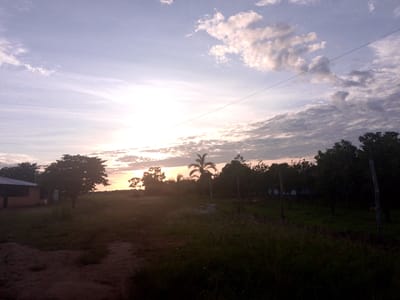 The image depicts an outdoor scene at dusk or dawn, featuring a sky with scattered clouds and a low-lying sun. In the foreground, a dirt path leads into a grassy area with several trees. To the left, a building with a dark roof and brown walls is partially visible. The overall environment suggests a rural or semi-rural setting, possibly within the municipality of Puerto Gaitán, Colombia, given the presence of palm trees and lush vegetation characteristic of the region. The lighting indicates a transition between day and night, casting long shadows across the landscape. No people are clearly discernible in the scene. The image appears to be captured with a handheld camera, as suggested by the slight blur and graininess.
