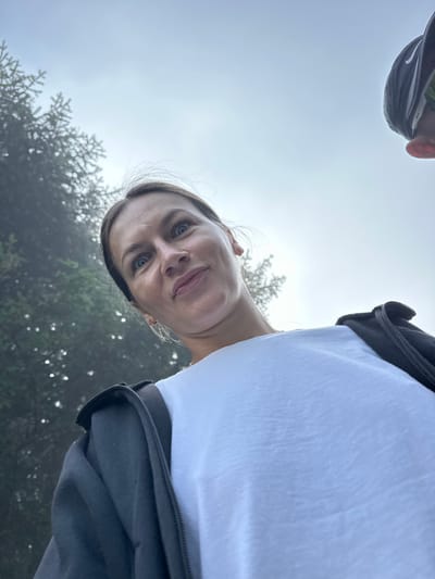 A low-angle, close-up, outdoor photograph captures a woman with light brown hair, her face turned towards the camera, eyes wide with a slight smirk. She is wearing a white t-shirt and a black zipped jacket. Behind her, blurred green foliage suggests a natural, possibly forested, environment. A portion of another person's head, wearing a black baseball cap, is visible in the upper right corner of the frame. The sky above is overcast and pale. This image appears to have been taken in Trešnjevik, Montenegro, given the context of the surrounding trees and the type of landscape typically found in that region.