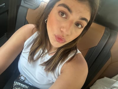 A close-up selfie-style image features a light-skinned female with brown hair. She is wearing a white tank top and a silver chain necklace. A black, textured bag is visible in the upper right corner of the frame. The background appears to be the interior of a car, with brown leather seats. Based solely on the image, it is not possible to pinpoint the specific location in Acapulco de Juárez, Mexico. There are no overt indications of activity, interaction, or environment that would allow for such localization within the city.