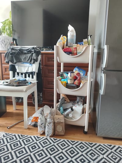 The image depicts an indoor scene, possibly a living room or dining area, with a collection of objects arranged on the floor, a chair, a multi-tiered cart, and a dresser. The overall impression is one of clutter. In the background, a large, dark television screen is visible, with a potted plant to its left. To the right of the television, a silver refrigerator stands.
In the foreground, a white rug with a black geometric diamond pattern covers a portion of the wooden floor. Placed on the rug are two large, silver-colored bags and one lighter-colored bag with Polish text that appears to be food packaging.
To the left of the rug, a white chair with a light-colored cushion is present. On the seat of the chair are various items, including what appears to be a blue item, possibly a tool, and some smaller objects. Draped over the back of the chair are several items of clothing, including t-shirts and what looks like athletic wear.
Next to the chair is a wooden dresser with multiple drawers, some of which are slightly ajar. On top of the dresser, more folded clothing is piled.
A white, multi-tiered rolling cart is situated between the dresser and the refrigerator. The cart is filled with various food items, including bags of groceries, jars, and what might be snacks. Some items are spilling out of the cart.
On the lower shelf of the cart, a metal grater and what looks like a small appliance are visible.
The lighting in the room appears to be artificial, casting soft shadows. The timestamp "01.07.2026 11:20" is superimposed on the lower right corner of the image.
The scene is located in Lębork, Poland.