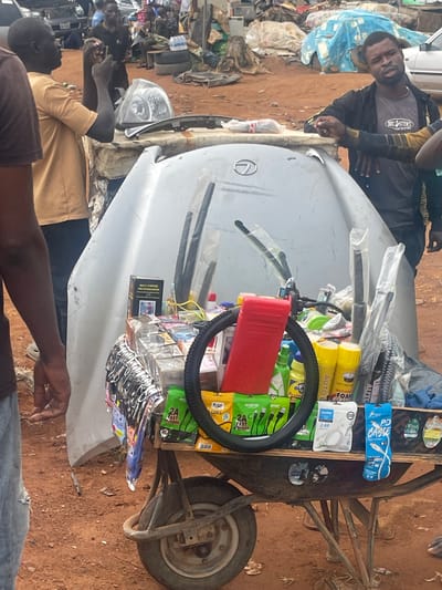 The image depicts a roadside scene in Karshi, Nigeria, featuring a hand-drawn cart used as a mobile stall. On top of the cart, covered with a white Lexus car hood, is a variety of goods for sale, including car accessories like windshield wipers, "AUTO FOAM" car care products, phone chargers (brands like "oraimo," "Wind," and generic 2.4A and PD cables), a bicycle tire, "MULTI-PURPOSE FIRE EXTINGUISHER," and packets of "FLICK" cables. Several men are standing around the cart. The surrounding environment appears to be somewhat littered, with debris visible in the background. The ground is reddish-brown, indicating the local soil composition.