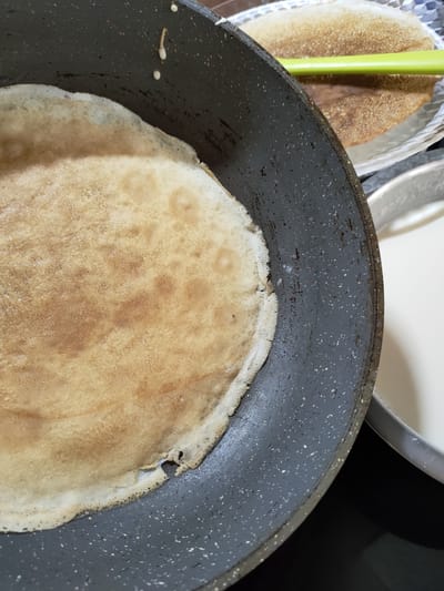 This is a close-up, top-down, eye-level shot of a crepe being cooked in a dark grey non-stick pan. The crepe is mostly cooked and has a golden-brown color with some lighter and darker patches. The edges of the crepe appear slightly crisp and are lifting from the pan. There are a few small bubbles on the surface of the crepe, and a small tear is visible near the edge. The pan has a speckled pattern of white and grey. To the right of the pan, a shallow silver dish contains a white liquid, likely batter. Behind the pan, another crepe is visible on a white plate, with a green spatula resting on it. The background is dark and out of focus. The scene appears to be set in a kitchen. There is no indication of the city of Perpignan, France.