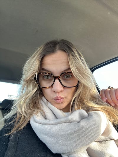 An individual with blonde hair and darker roots, wearing large, tortoise-shell framed eyeglasses, is depicted in a selfie captured from within a vehicle. The subject's light green/hazel eyes are visible through the lenses, and their lips are puckered. A light beige knitted scarf is wrapped around their neck, partially obscuring a dark outer garment. A gold-toned ring is present on the ring finger of the left hand, visible on the right side of the frame. The vehicle's gray fabric headliner is visible above the subject, and a bright outdoor light source is discernible through a window on the right. A car seat headrest is partially visible in the background on the left. The scene is located in the city of Veauche, France.