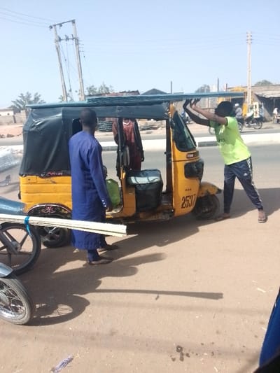 The image depicts a daytime outdoor scene at a roadside location near Muhammadu Dikko Stadium, Nigeria. The central focus is a stationary yellow three-wheeled auto-rickshaw (keke napep) with a black canvas roof, marked "KTN" and "2537" on its side. A stack of corrugated metal sheets is loaded horizontally onto the auto-rickshaw's roof.
Three individuals are visible. On the right, a male wearing a light green t-shirt and dark blue athletic pants with white stripes is actively pushing upwards on the corrugated sheets with both hands, appearing to adjust or secure them. To the left, a second male, wearing a dark blue long-sleeved garment, stands with his back mostly to the viewer, observing the scene. A third individual, partially obscured and wearing a dark red garment, is visible inside or immediately behind the auto-rickshaw's passenger compartment. Inside the auto-rickshaw, a light green bag and a dark insulated container are present.
In the immediate foreground on the left, several long, thin, light-colored rods are secured with blue string to the side of a motorcycle, of which only the front wheel and part of the frame are visible. The auto-rickshaw is positioned on a dirt or gravel shoulder adjacent to a paved road, which features a white curb line. The background includes multiple utility poles and power lines, one noticeably tilted, against a clear, light blue sky. Distant buildings and sparse vegetation are visible. Strong overhead sunlight casts distinct shadows of the subjects onto the ground. The scene portrays the loading or securing of building materials onto transport vehicles.