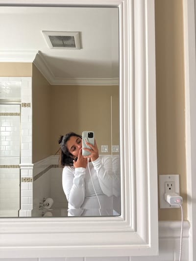 The image depicts a mirror selfie taken by a female subject in a private bathroom located in Princeton, United States. The subject, visible from the chest up in the mirror's reflection, has dark hair partially tied back and wears a white long-sleeved top. Her left index finger is positioned near her lips in a gestural pose. Her right hand holds a light-colored smartphone, which has a red/pink accessory attached to its back. A white braided charging cable extends from the phone to a white wall charger, which is plugged into a standard white duplex electrical outlet on the beige wall immediately adjacent to the mirror's white ornate frame. The charger emits a pink indicator light. The reflected bathroom interior features white subway tiles with a decorative brown and white accent border, beige painted walls, white crown molding, a rectangular white ceiling vent, and a white toilet paper holder with a roll.