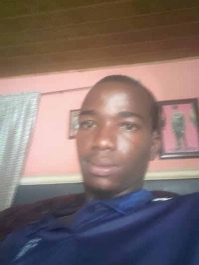 The image is a close-up, blurry selfie of a young Black male wearing a navy blue polo shirt. He is positioned in the center of the frame, with his face slightly turned to the right. The background consists of a pale pink wall, a framed picture on the wall depicting a person in uniform, and a section of a wooden slatted ceiling. To the left of the frame, a light-colored, textured curtain is visible. The lighting appears to be from an overhead source, casting highlights on the subject's face. The overall impression is of an indoor setting, likely a residential or personal space. Without further context or visible landmarks, it is not possible to pinpoint the exact location within Oturkpo, Nigeria.