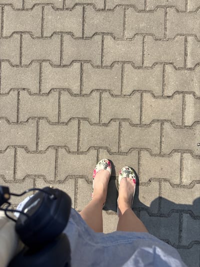 This media file shows a close-up, overhead view of a person's legs and feet standing on a grey, interlocking paver pathway. The pavers have a distinct 'I' shape. The person is wearing light-colored flat shoes adorned with a floral pattern featuring pink roses and green leaves, along with a small, silver orb embellishment. A portion of a light blue garment, possibly a dress or skirt, is visible on the right side of the frame.
In the lower right corner, a pair of black headphones is visible, resting on a light-colored surface, possibly a bag or stroller. The lighting suggests it is daytime, with distinct shadows cast on the pathway, indicating a sunny environment. The scene appears to be outdoors, likely in a public space like a park or a sidewalk. The overall impression is one of casual leisure or a moment of pause during an outdoor activity. No visible text is present in the image. The location context provided indicates Červená Píska, Czechia.