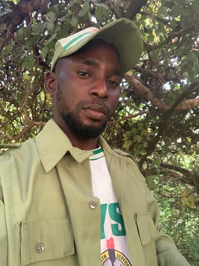 A dark-skinned adult male is depicted in a close-up, chest-up portrait, looking directly at the viewer with a neutral expression. He has short dark hair and a well-trimmed beard. He is wearing a light olive-green long-sleeved shirt or jacket with shoulder epaulets and two buttoned chest pockets. Underneath, a white t-shirt with green stripes on the collar and a partial graphic, including the letters "YS" in green and a logo with a torch and "YOUTH SERVICE" text, is visible. An olive-green baseball cap, featuring white and green stripes on the brim and a small red and white emblem, is worn on his head. The background is a dense, blurred composition of green foliage and tree branches, indicating an outdoor, natural setting. The specific location within the city of Kubi A, Nigeria, is not visually identifiable in the image.