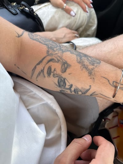 The image shows a close-up of a person's arm with a tattoo. The tattoo appears to be a stylized face or portrait with intricate details. The arm is positioned in front of a person wearing light-colored clothing and a black purse is visible in the background. Another person's hands are also visible in the foreground. The scene suggests an indoor setting, possibly a car or a public transport, given the clothing and the presence of a bag. There are no identifiable landmarks or elements that would place this image specifically in the city of Proshyan, Armenia.