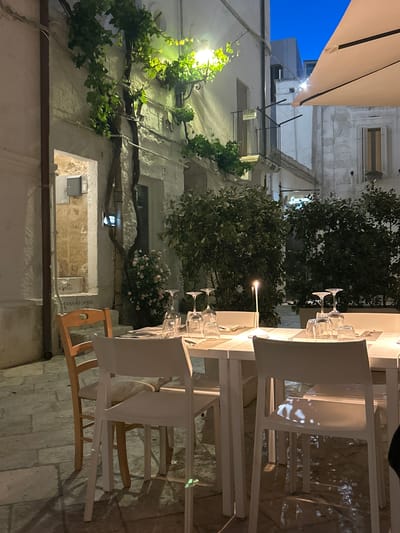 The image shows an outdoor dining scene in Polignano a Mare, Italy, at night. A white tablecloth-covered table is set with glassware. White plastic chairs are placed around the table, one of which has a wooden backrest. A lit candle is placed on the table. Green plants and trees surround the dining area. The building walls appear to be made of white stone or stucco. The sky above is dark blue, seen partially from under a sunshade. A grape vine runs along the side of the building.