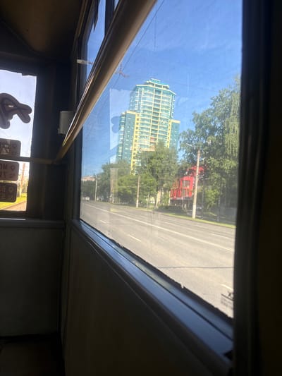 A view from inside a moving vehicle, likely a bus or tram, looking out a window. The window frame is dark, and a yellow handrail is visible along the top. Outside, a street runs parallel to the direction of travel. On the right side of the street, a tall, modern apartment building with a turquoise and yellow facade stands out. Further down the street, a red building with a modern design is visible. Trees line the street, and the sky is clear and blue. On the left side of the interior, a window shows a sign with some Cyrillic text and icons, indicating a bus stop or route information. The overall scene suggests an urban environment in Kirov, Russia, on a sunny day.