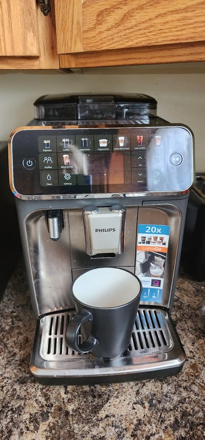 This is a head-on, medium shot of a Philips espresso machine, positioned on a speckled brown countertop. Above the machine, wooden kitchen cabinets are visible. The espresso machine features a digital display screen showing various coffee options and settings. Below the screen, the Philips logo is displayed on a chrome panel. A black mug sits on the drip tray of the machine, ready to receive coffee. A small, blue and white advertisement for "LatteGo" is affixed to the right side of the machine. There are no people present in the image. The scene is indoors, likely in a kitchen setting. The precise location in St. John's, Canada, cannot be determined from the image alone.