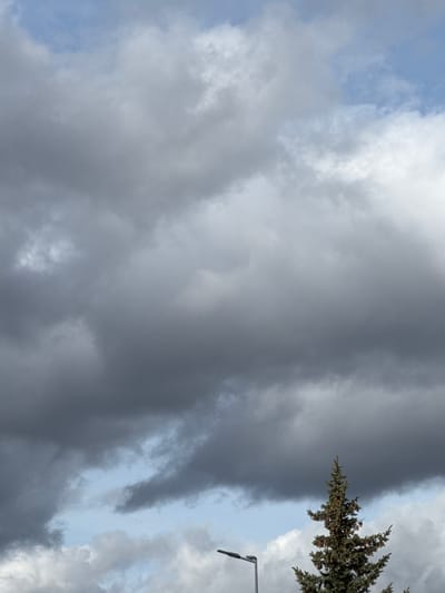 The image captures a dynamic sky dominated by clouds, with portions of a tree and a lamppost visible in the upper right quadrant. The perspective suggests the photograph was taken looking upwards towards the sky.
The sky is a mix of heavy, dark grey clouds and brighter, whiter cloud formations. A large mass of dense, dark grey clouds spans across the left and central parts of the frame, suggesting an overcast or approaching stormy condition. Interspersed with these darker clouds are areas of lighter, fluffier white clouds, particularly noticeable in the upper left and middle right areas of the sky. Patches of clear, light blue sky are visible, most prominently in the upper left corner, providing a contrast to the dense cloud cover.
In the upper right corner of the image, the top branches of a coniferous tree are visible. Its dark green foliage, possibly consisting of pine or spruce needles, extends into the frame from the right edge. The tree appears full and healthy.
Immediately below the tree, also on the right side of the frame, is a partial view of a metallic lamppost. Only the upper arm of the post and a portion of its light fixture or attached sensor are discernible. The lamppost is dark in color and contrasts against the lighter sky and clouds behind it.
There is no specific activity taking place, and no visible text in the image. The scene depicts an outdoor setting under varying cloud conditions, possibly indicating changing weather or a transient moment during the daytime.