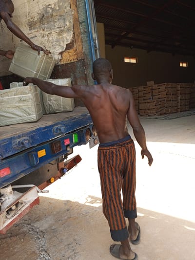 A man with a bare upper torso and wearing geometrically patterned pants and sandals is standing near the rear of a truck while other men are loading large, wrapped packages onto the truck. The truck is parked inside a warehouse or storage facility filled with stacks of boxes labeled "NASOR". The scene is brightly lit, suggesting daytime. The location is likely in Pasere, Nigeria, as requested.
