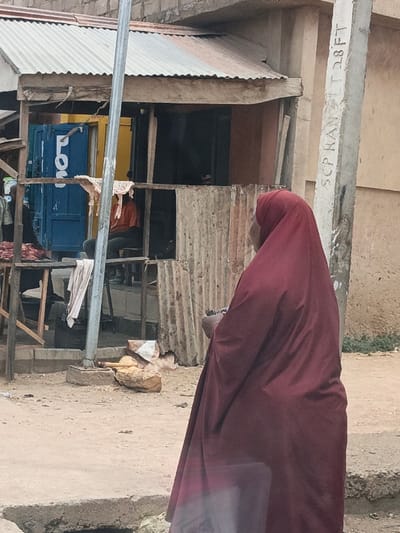 The scene is an outdoor urban environment in Hotoro South, Nigeria, depicting a street adjacent to commercial structures. In the immediate foreground, a person clad in a full-length, deep maroon garment stands with their back to the observer, facing towards the mid-ground. Their left hand appears to hold a small, dark object.
A concrete utility pole stands vertically in the mid-ground to the right, bearing faded white vertical text "SCP KAN T 28FT." A slender metal pole is positioned slightly left of center.
In the mid-ground, a makeshift market stall or storefront is visible. It features a corrugated metal roof over an open area. The structure is built with a wooden framework and corrugated metal sheets. A blue door, partially open to reveal a yellow interior wall, has the partial white lettering "LO" on its exterior. Inside this structure, a person wearing an orange-red shirt and dark trousers sits on a stool. To the left of this individual, behind a counter, another dark-clothed figure is partially obscured, near what appears to be cuts of raw red meat. A light-colored cloth is draped over a horizontal beam within the stall.
The ground is packed dirt. A concrete curb and an open drainage channel are present in the lower left. A wooden pestle-like object and fragmented concrete or stone pieces lie on the dirt ground near the base of the metal pole. A light orange/peach building wall forms the background behind the market stall. Some sparse greenery is visible in the far background on the right. The lighting is overcast.
