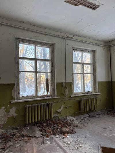 Two adjacent windows, one above the other, look onto bare trees in an abandoned building. Paint peels from the walls, which transition from white to green halfway down. Radiators hang next to each window. The floor is covered with debris. A section of lattice is missing from the wall above the windows. The building appears derelict and in disrepair.