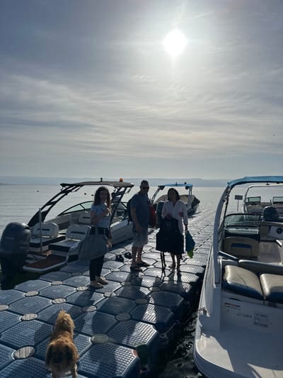 The image shows three adults standing on a floating dock at the Sea of Galilee in Migdal, Israel (latitude: 32.844, longitude: 35.52). The adults are standing in the middle of the dock with two boats on either side of them. The people appear to be in their 30s and 40s. The woman in the middle is wearing a black jacket and black pants. The woman on the left is wearing a long, dark-colored dress and a black shirt. The man on the right is wearing a gray t-shirt and black pants. They are all looking toward the camera, and it appears as though they have just gotten off the boats. The boat on the left is a black speedboat with a white deck. The boat on the right is a white speedboat with a gray and white deck. There is a brown dog sitting in the middle of the dock with a white shirt on. The sun is shining brightly in the sky, and there is a bright blue sky with white clouds behind the people. There is blue water in the background and some mountains in the distance. There are shadows from the people on the floating dock behind them. The scene is calm and peaceful.