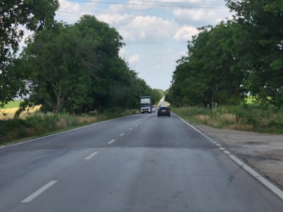 This media file captures a view from a vehicle traveling on a paved road through a rural, tree-lined landscape. The road is asphalt, marked with white dashed lines indicating lanes. A few cars and a truck are visible on the road ahead, suggesting moderate traffic. The scene is flanked by dense green trees on both sides, with glimpses of fields, possibly agricultural, in the distance. The sky is partly cloudy with white cumulus clouds against a bright blue backdrop, indicating fair weather. The lighting suggests it is daytime, likely late morning or early afternoon, due to the brightness. There is no visible text in the image. The overall impression is one of a pleasant, ordinary journey through the Bulgarian countryside near Zgalevo.