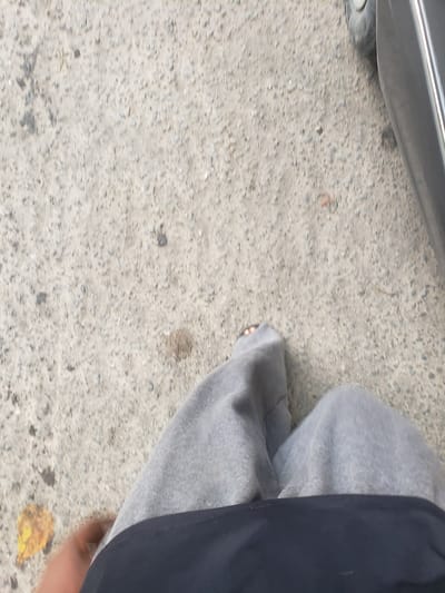 The image shows a first-person perspective of a person standing on a textured concrete surface, possibly a sidewalk or road. The person is wearing grey sweatpants and a dark blue garment. One bare foot with visible toes is on the ground, with a yellow leaf nearby. To the right, the lower portion of a dark-colored vehicle, including a tire and part of the body, is visible. The environment appears to be outdoors, in an urban setting. The concrete surface is speckled with small stones and debris.