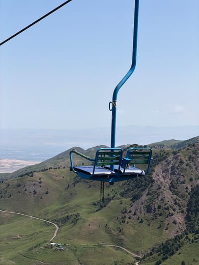 A blue ski lift chair numbered "37" hangs suspended against a clear, pale blue sky, its seat made of blue metal and wooden slats. The chair is attached to a thick black cable that extends diagonally upwards and to the left. Below, a lush green mountainous landscape unfolds, characterized by rolling hills dotted with scattered trees and shrubs. A winding dirt road snakes through the valley, leading to a cluster of buildings with light-colored roofs. In the distance, a hazy, pale landscape stretches to the horizon. The scene appears to be the city of Kashka Suu, Kyrgyzstan.