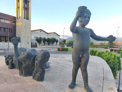 This media file showcases an outdoor scene with two sculptures in the foreground. To the right, a bronze statue of a young boy stands holding a chalice-like object in his raised right hand and a bunch of grapes in his left arm. The boy is depicted nude. To the left, a larger, abstract bronze sculpture appears to represent a group of animals or figures in a more organic, flowing form. The sculptures are situated on a paved area.
In the background, a modern, light-colored building with a tower and dark-paneled sections stands on the left. Adjacent to it is another building with a lighter facade and orange-tiled roofs, featuring signage that reads "La Rioja Alavesa" and "BODEGAS VINAS ARANA". A street with a roundabout is visible in front of these buildings, with a red circular sign indicating "no entry". Greenery, including trees and vines, surrounds the buildings and extends into the distance, suggesting a vineyard or agricultural setting. Mountains can be seen on the horizon. The sky is clear and blue, indicating it is daytime, likely late afternoon or early morning given the lighting. The overall atmosphere is peaceful and hints at a rural or semi-rural location associated with wine production in Haro, Spain.