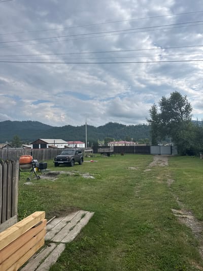 The image shows a rural yard in Yabogan, Russia, on a cloudy day. In the foreground, stacks of new lumber rest on weathered wooden planks. To the left, a wooden fence borders the property. In the middle ground, a dark-colored Lada Niva SUV is parked on the grass. Near the vehicle, an orange cement mixer sits next to a black container. A dirt path leads towards a metal gate in the background. Beyond the gate, a cluster of buildings with peaked roofs is visible. The yard is covered in green grass, and a dirt track runs through it. In the distance, rolling, forested hills rise under a sky filled with dense, grey clouds, suggesting an overcast or approaching storm. Power lines are visible across the top of the frame.