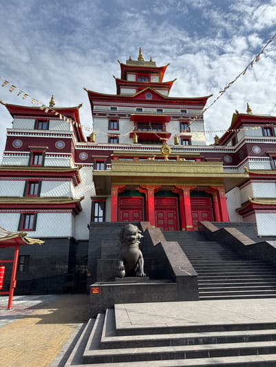 The image depicts a tall, multi-tiered Buddhist temple in Kyzyl, Russia, characterized by a mix of white, red, and gold architectural elements. The temple's facade is adorned with windows and decorative patterns. To the right of the temple is a dark gray, monolithic structure with steps leading up to it, and a lion statue at its base. The sky is visible in the background, with white clouds and colorful prayer flags strung between the temple structures. The ground near the bottom right of the frame is paved with bricks.