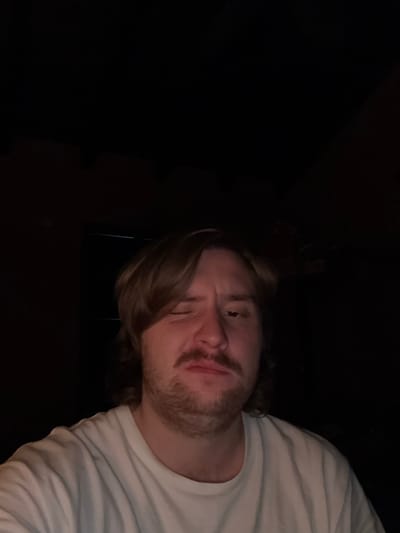 A man with light brown hair and a beard is visible from the chest up, wearing a white t-shirt. The background is mostly dark, with some indiscernible features. The photograph appears to be taken from a low angle, looking up at the man. No identifiable elements link the image to Cañada de Madero, Mexico.