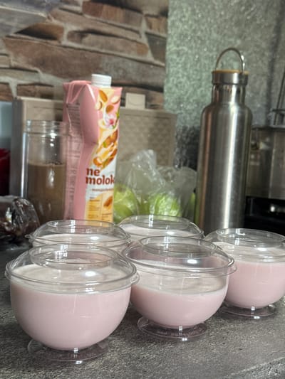 The image depicts a counter or shelf in what appears to be a kitchen or food preparation area. There are several items present: a stainless-steel water bottle, a carton of "ne molok" almond milk, possibly green apples in a clear plastic bag, some jars/containers, and four clear plastic cups filled with a pink liquid, possibly a yogurt or smoothie. The background includes a brick-like patterned wall. It is not possible to verify the location in Kurganinsk, Russia.