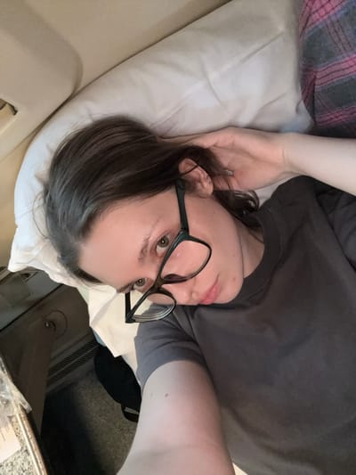 This is a close-up, over-the-shoulder selfie taken indoors. The subject is a person lying on their side, propped up by a white pillow. They are wearing a dark gray t-shirt and what appears to be a pink and gray plaid blanket is visible in the upper left corner of the frame. The person has dark hair with some gray streaks, is wearing black-rimmed glasses, and has piercings in their left ear. Their right arm is bent and their hand is resting near their ear and hair. The person's face is turned towards the camera, and they appear to be looking directly at it with a neutral expression.
The setting seems to be a room, possibly a bedroom or a hospital room, given the pillow and the faint outline of what might be a hospital bed rail on the left. In the background, there is a dark surface with a patterned texture, possibly a floor or furniture, and some text on a light-colored paper can be seen in the lower right corner, though it is largely illegible. The lighting suggests it is daytime, but there are no direct cues about the weather. The image is oriented sideways, with the subject's head towards the bottom right of the frame.