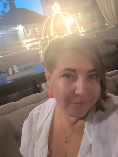 A woman is taking a selfie in what appears to be an outdoor seating area of a restaurant. She is wearing a white open-collared shirt and large pink hoop earrings. A gold necklace with a cross pendant is visible. She has short brown hair and is looking at the camera. In the background, there are upholstered seats, tables, and decorative lights. The setting is likely an evening or nighttime outdoor dining establishment in Alfimovo, Russia.
