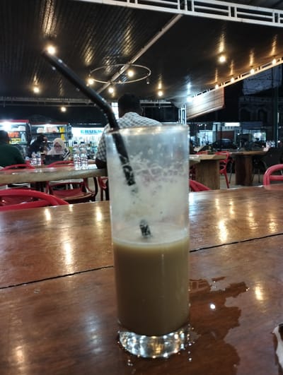In a dimly lit outdoor seating area, likely at a restaurant or cafe in Blangmetimu, Indonesia, a glass of iced coffee sits on a wooden table. The beverage is light brown with a foamy top and a black straw. The table surface is wet, with reflections of overhead lights visible. In the background, several people are seated at other tables, some under a corrugated metal roof structure illuminated by string lights. A woman wearing a hijab is visible at one table, looking at a laptop. The overall atmosphere suggests a casual evening gathering.