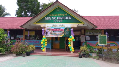 The image shows the entrance to MIN 50 Bireuen, a school located in Julicotmeurak, Indonesia. A red tiled roof covers the entrance area, supported by pillars. A green banner above the entrance reads "Selamat datang di MIN 50 BIREUEN ACEH" (Welcome to MIN 50 Bireuen Aceh). Below this, a banner announces "HARI PERTAMA MADRASAH BERSAMA AYAH" (First Day of Madrasah with Father). Colorful balloons in yellow and green are arranged on either side of the entrance. To the left, potted plants and bushes are visible, with a few individuals in the background, including a woman wearing a hijab. To the right, more potted plants and a colorful mural depicting children and nature are present. Several signs with text in Indonesian are posted on the building. The ground in front of the entrance is paved with green and white checkered tiles. Two men are visible standing at the entrance.