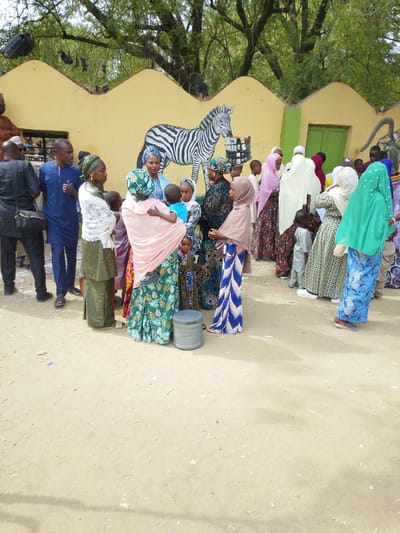 The image shows a group of people gathered in what appears to be a public space in Garun Gawa, Nigeria. Many of the women are wearing traditional Muslim attire including headscarves. Some are holding children. Men are also present, dressed in both traditional and western clothing. In the background, there is a low building with a mural of a zebra and other animals. The ground is dirt or sand, suggesting an outdoor environment. The presence of trees and painted murals indicates the scene may be near a school or park. The general atmosphere seems relaxed and informal.