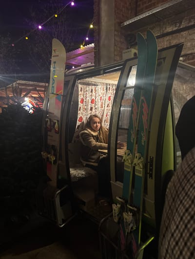A woman with long blonde hair and a tan jacket lies on a cushion with a beige pattern under what appears to be stacked canoes and skis. Two pairs of light blue skis with red and black text are on top, while a pair of white HEAD skis with pink, light blue, and yellow markings are on the bottom. Colored string lights hang in the darkness nearby.
