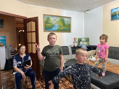 A residential interior in Bely Klyuch, Russia, features four individuals: an elderly woman seated on a small stool holding a tablet; a young boy holding up a peace sign; a younger boy with arms outstretched; and a young girl standing on a couch. The environment includes a door frame, paintings on the wall, and a couch. The elderly woman is dressed in a dark floral-patterned top and dark slacks. The children are in casual attire. The space is decorated with framed landscape paintings and dolls on a shelf.