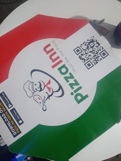 A pizza box from Pizza Inn features a white background with red and green curved sections at the top and bottom, respectively. The "Pizza Inn" logo is prominently displayed in green and red, accompanied by the tagline "Must be the Pizza". A cartoon chef character is included. A QR code is placed near the top edge. The box also includes download options for the Pizza Inn Kenya delivery app from the Google Play Store and the Apple App Store.